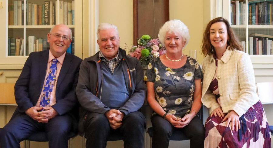 Dr. Kevin Whelan, Fr. Peter McVerry, Judy Friel, and Eimear Clowry Delaney sitting, smiling in front of one of Friel's beloved grandfather clocks donated by Anne Friel to accompany the Brian Friel Library collection at O'Connell House.