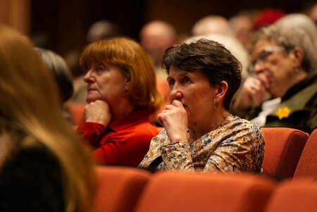 Two women listen intently in an auditorium. The woman in the foreground, wearing a light patterned shirt, rests her chin on her hand. The woman next to her, wearing a bright red shirt, also has her chin resting in her hand.