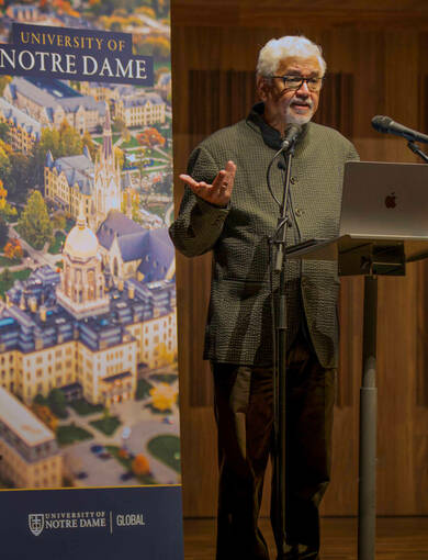 A man gestures with his right hand while speaking at a podium. He wears a textured dark green blazer and brown pants. Behind him, a banner displays an aerial view of the University of Notre Dame campus, including the Main Building's Golden Dome, along with the University of Notre Dame Global logo. A laptop and two microphones are positioned on the podium.