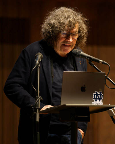 Stephen Rea, a man with curly gray hair and glasses stands at a podium, looking down at a laptop with an Apple logo and a Notre Dame Research sticker. Two microphones are positioned in front of the speaker.