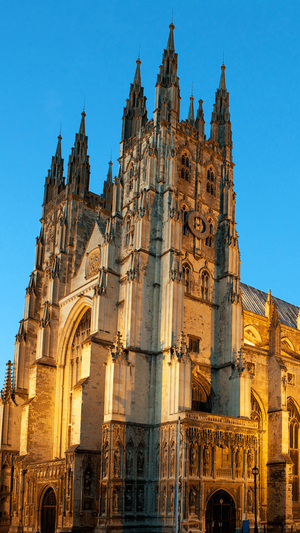 Canterbury Cathedral's ornate, tan stone facade and towering spires at dusk. The clock face is visible, and warm light illuminates the detailed carvings near the entrance.