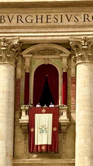 Pope on a balcony of St. Peter's Basilica with a maroon and white Papal banner hanging below.