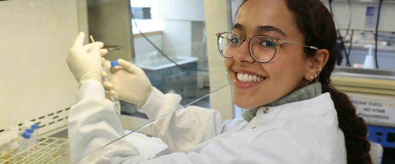 A smiling young woman in a white lab coat and glasses looks at the camera while holding a blue-capped vial with gloved hands inside a lab hood.