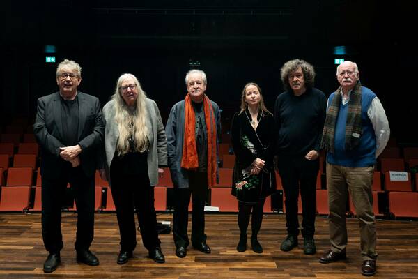 Four men and two women stand on a wooden stage before rows of red theater seats. They include a man in a black suit, a woman with long gray hair, a man with an orange scarf, a woman in an embroidered black robe, a man with curly hair, and a man in a blue vest with a plaid scarf.