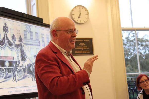 Bald man in red jacket and glasses smiles, gesturing while speaking at O'Connell House. Behind him, a screen displays "The Funeral of Daniel O'Connell" illustration. A wall plaque names Mary McAleese and Rev. Edward Malloy. A clock reads 7:00.