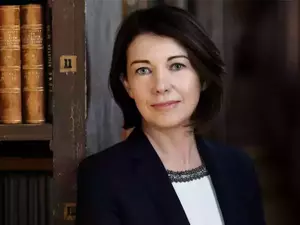 Prof. Linda Hogan. A woman with short dark hair and blue eyes smiles gently, wearing a navy blazer and white top with a beaded neckline, standing beside an old wooden bookshelf filled with brown leather-bound books.