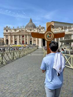 A person in a blue shirt, with a white jacket over their shoulder, views St. Peter's Basilica in Vatican City. A large wooden cross with a colorful Jubilee 2025 emblem stands in the foreground, with a crowd gathered in St. Peter's Square.
