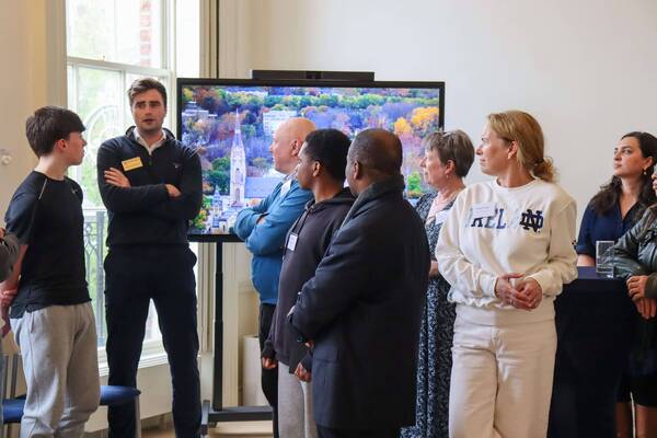 A group of attentive people, including a woman in a white Notre Dame sweatshirt, stand indoors. A large screen behind them shows an aerial view of the Basilica of the Sacred Heart and Notre Dame campus with colorful autumn trees.