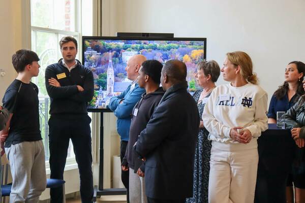 A group of attentive people, including a woman in a white Notre Dame sweatshirt, stand indoors. A large screen behind them shows an aerial view of the Basilica of the Sacred Heart and Notre Dame campus with colorful autumn trees.