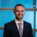A man with short dark hair and a beard, smiling and looking forward, wears a navy suit jacket, white shirt, and purple patterned tie with a silver lapel pin against a blurry blue background.