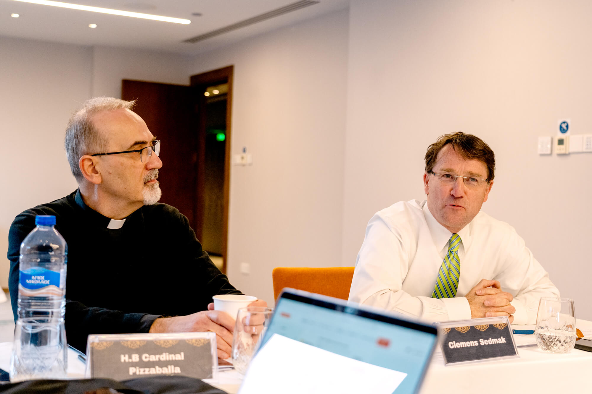 H.B Cardinal Pizzaballa in clerical attire looks right. Clemens Sedmak, wearing a white shirt and green striped tie, looks forward with a serious expression, hands clasped at a conference table.