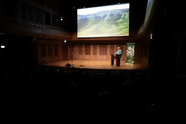 Man in blue jacket speaks at a podium on an auditorium stage, with a Notre Dame banner. Screen shows lush green terraced hills.