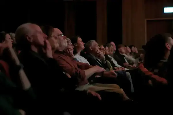 Attendees watch a presentation in a dimly lit auditorium. Several people rest their chins on hands, showing focused engagement.