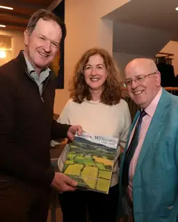 Three smiling people hold a book, "Gaelic Wexford," with a green landscape cover. One wears brown, one cream, one a blue suit.