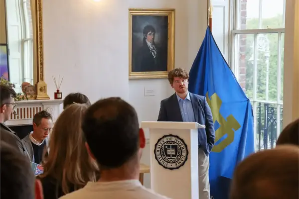 A man with curly red hair and a beard speaks at a podium displaying the University of Notre Dame seal. A blue Notre Dame flag is behind him.