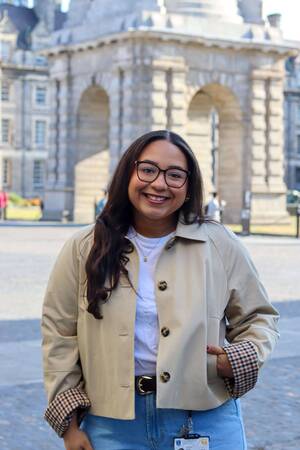 Smiling woman in glasses, beige jacket with plaid cuffs, stands before historic stone arches at Trinity College Dublin.