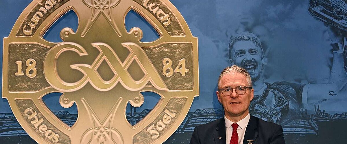 Man in suit and tie stands by a large gold Gaelic Athletic Association logo with "18 84" and text.