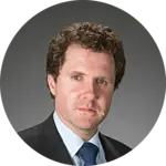 A man with curly brown hair and blue eyes wears a suit and blue tie.